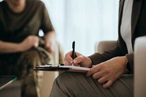 a military person sits in the background as their therapist writes down information on a clipboard during the military members veterans program
