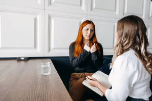 a woman with red hair sits at table with a woman with a notebook and is discussing how her anxiety treatment program has helped with her anxiety symptoms