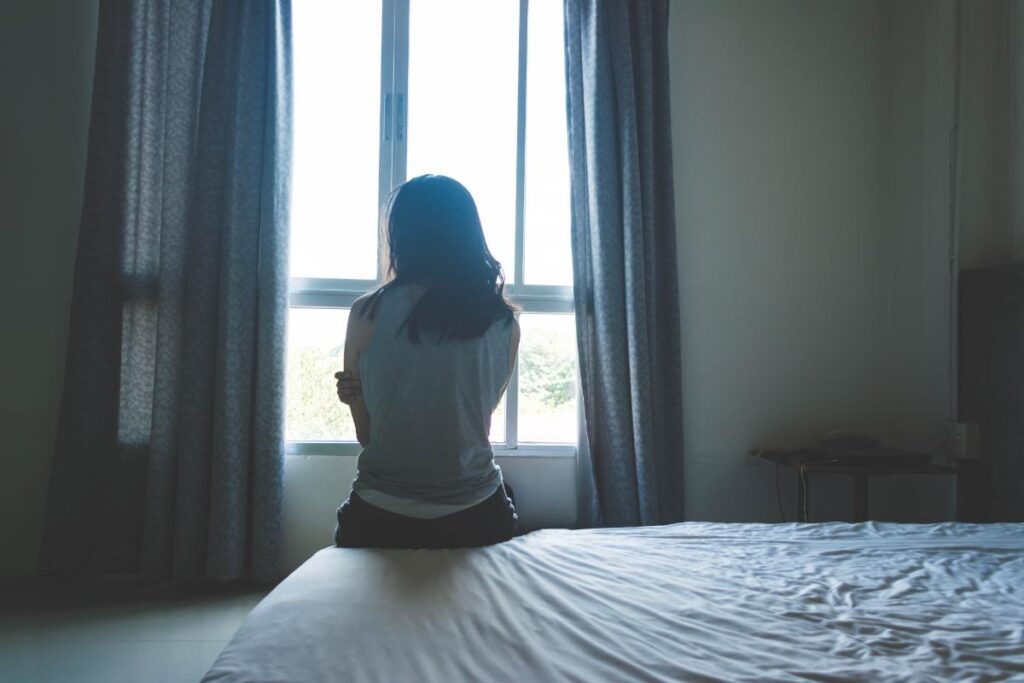 a person sits on the edge of bed looking out the window and experiencing the effects of alcohol and trauma
