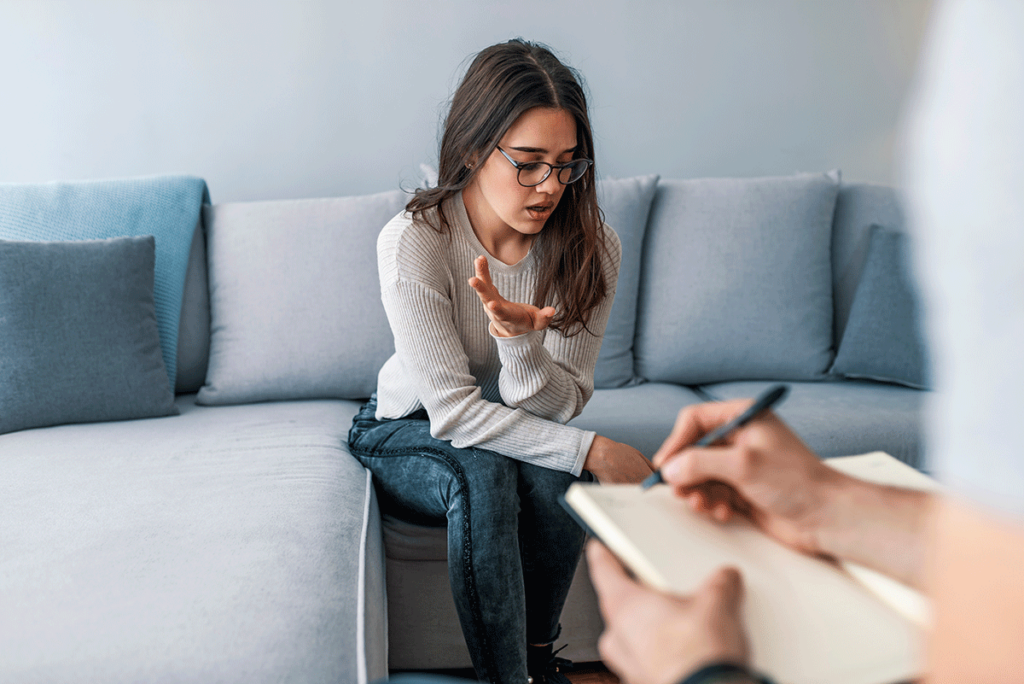 woman with glasses sits on a couch while she lists off the benefits or residential treatment to her therapist who is taking notes